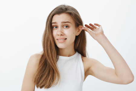 Stop lying my ears tired. Portrait of funny attractive woman with fair hair, pulling ear with hand and staring displeased at camera, feeling awkward while standing against gray background. Body language concept
