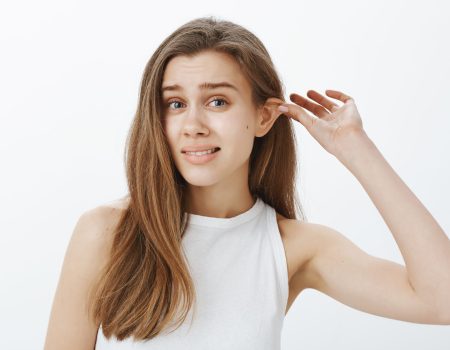 Stop lying my ears tired. Portrait of funny attractive woman with fair hair, pulling ear with hand and staring displeased at camera, feeling awkward while standing against gray background. Body language concept